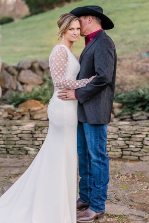 Couple in western wedding attire embrace next to stone wall during outdoor photoshoot.