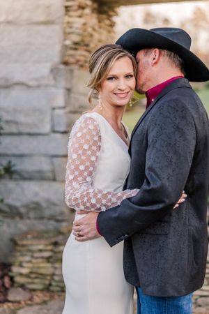 Romantic wedding portrait with couple in western attire against stone wall backdrop.