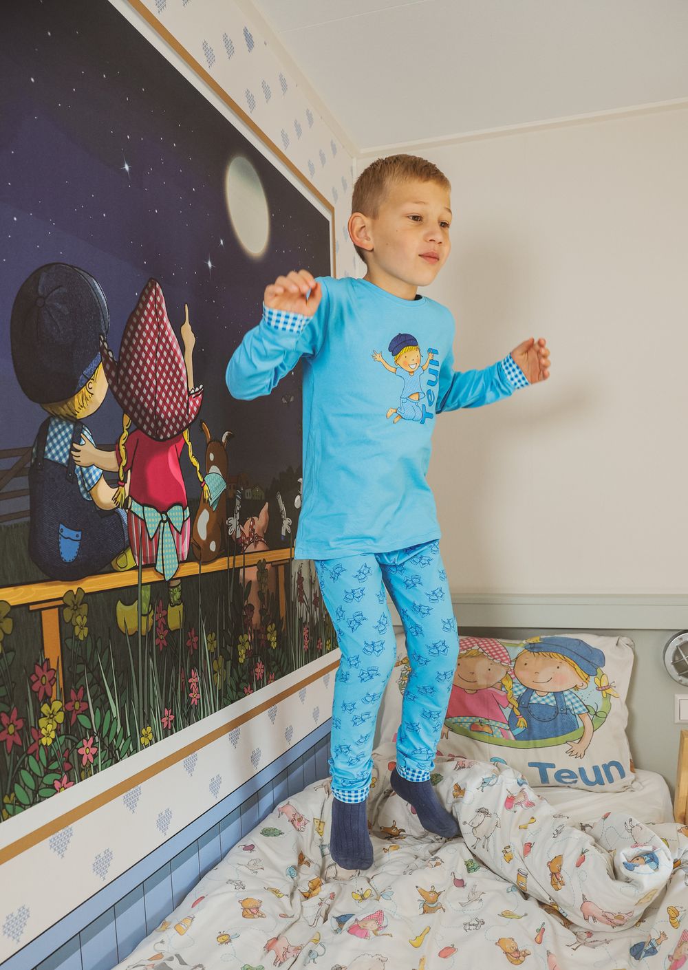 Child in light blue pajama set standing on bed against wall with colorful mural.