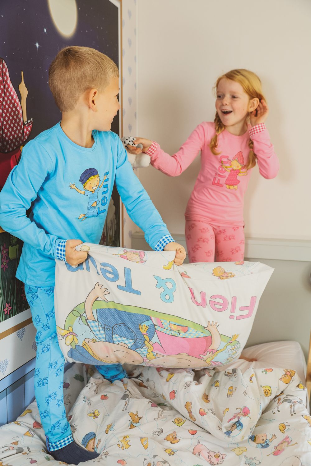 Two children in coordinating sleepwear playing with bedding on mattress.