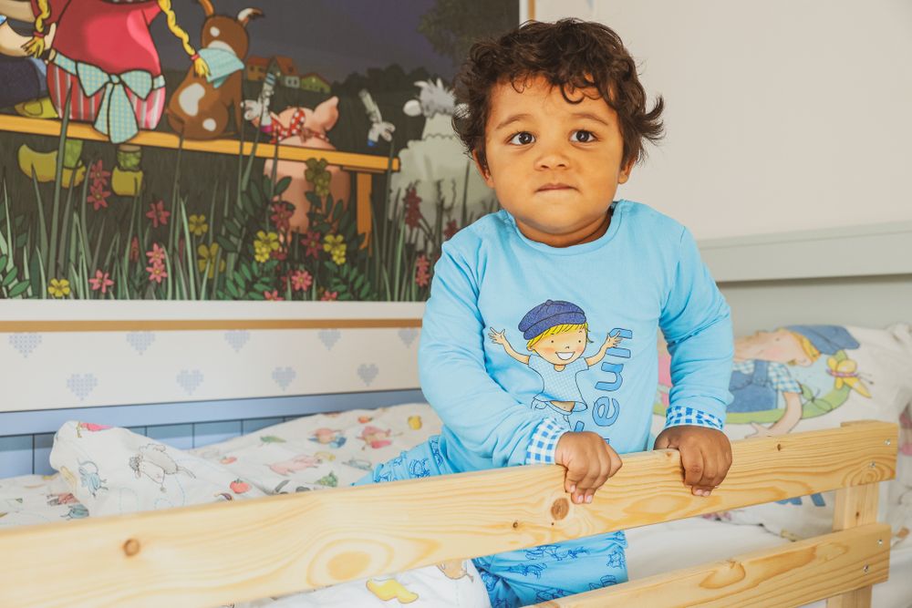A toddler in a light blue shirt stands in a bed with colorful bedding and wall decorations.