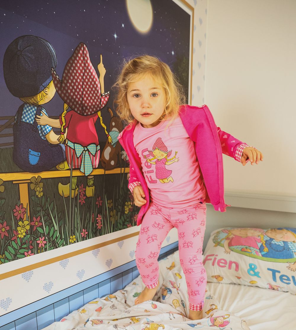 A child jumping on a bed wearing pink pajamas against a wall with whimsical nighttime artwork.