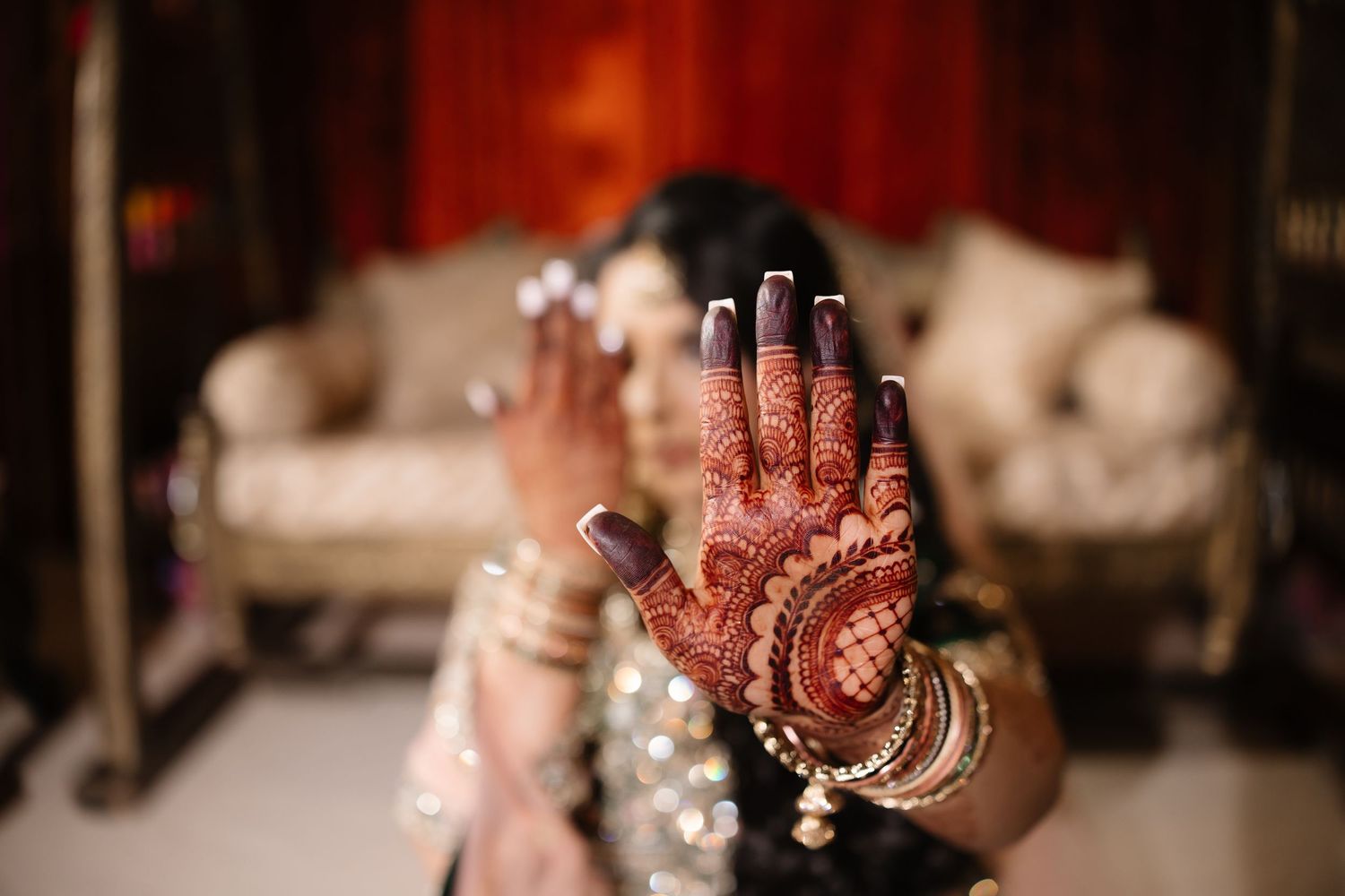 Two hands clasped together, one adorned with intricate henna designs and bangles against a red background.