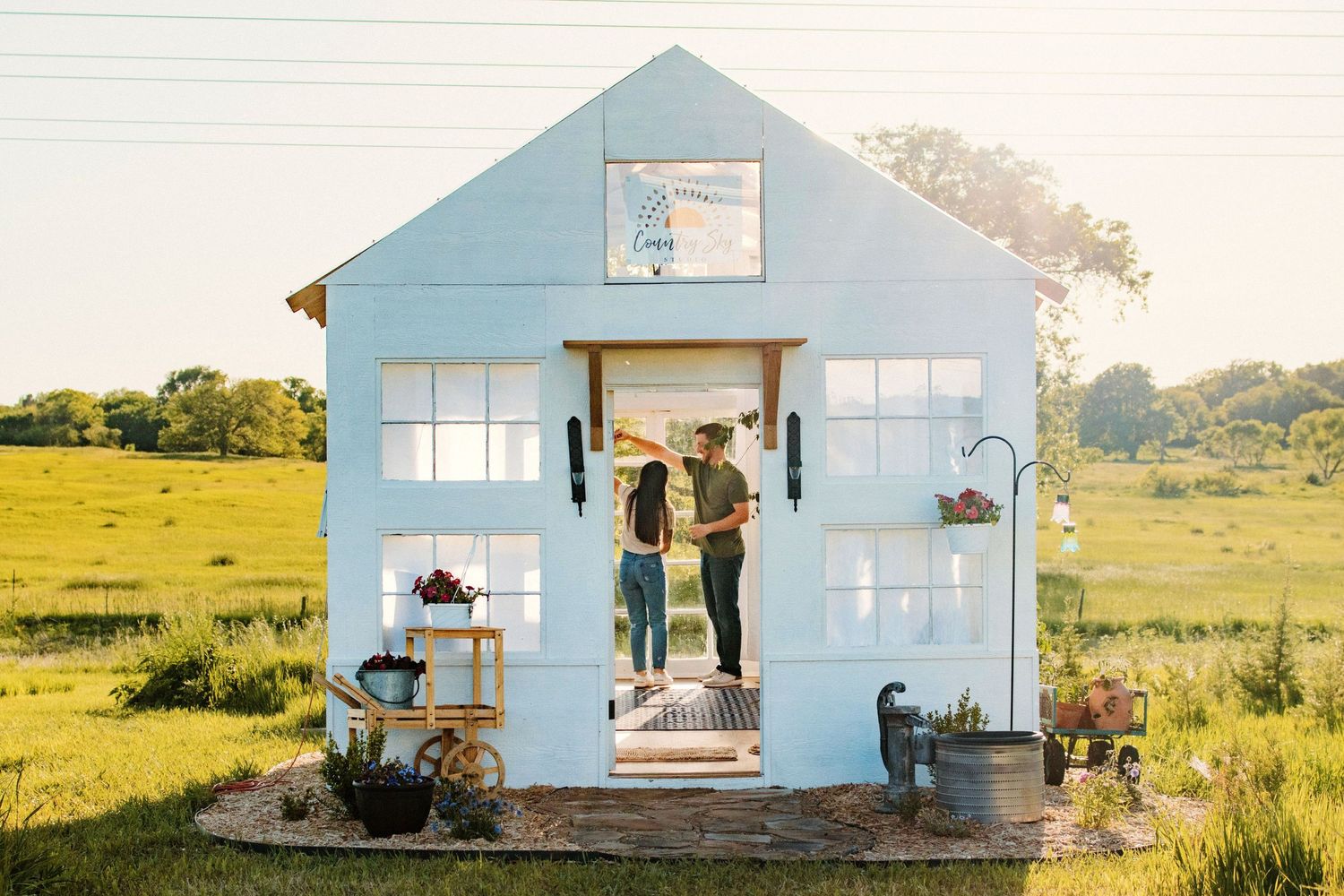 Couple standing in doorway of light blue tiny house in sunny meadow.