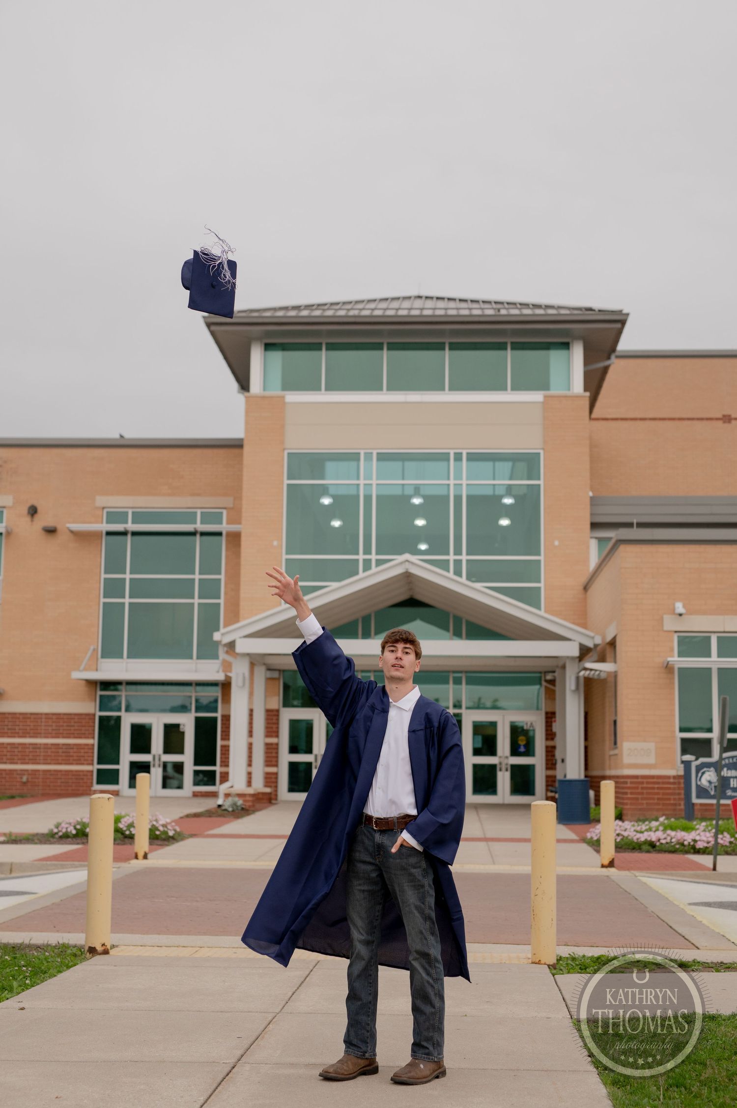 Student tosses graduation cap in celebration outside tan brick school building with glass entrance.