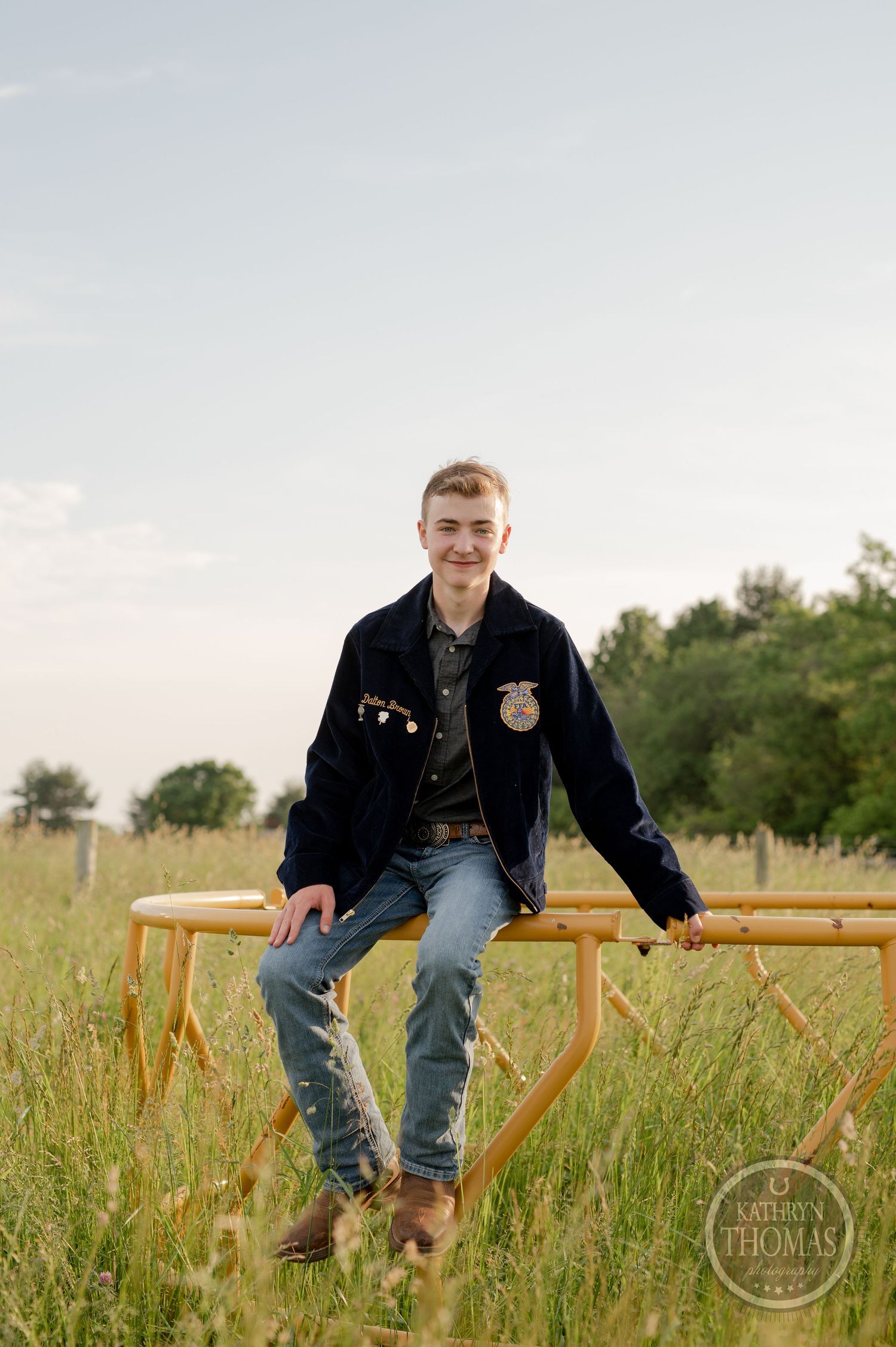 Student wearing FFA jacket sits on yellow barrier in grassy field.
