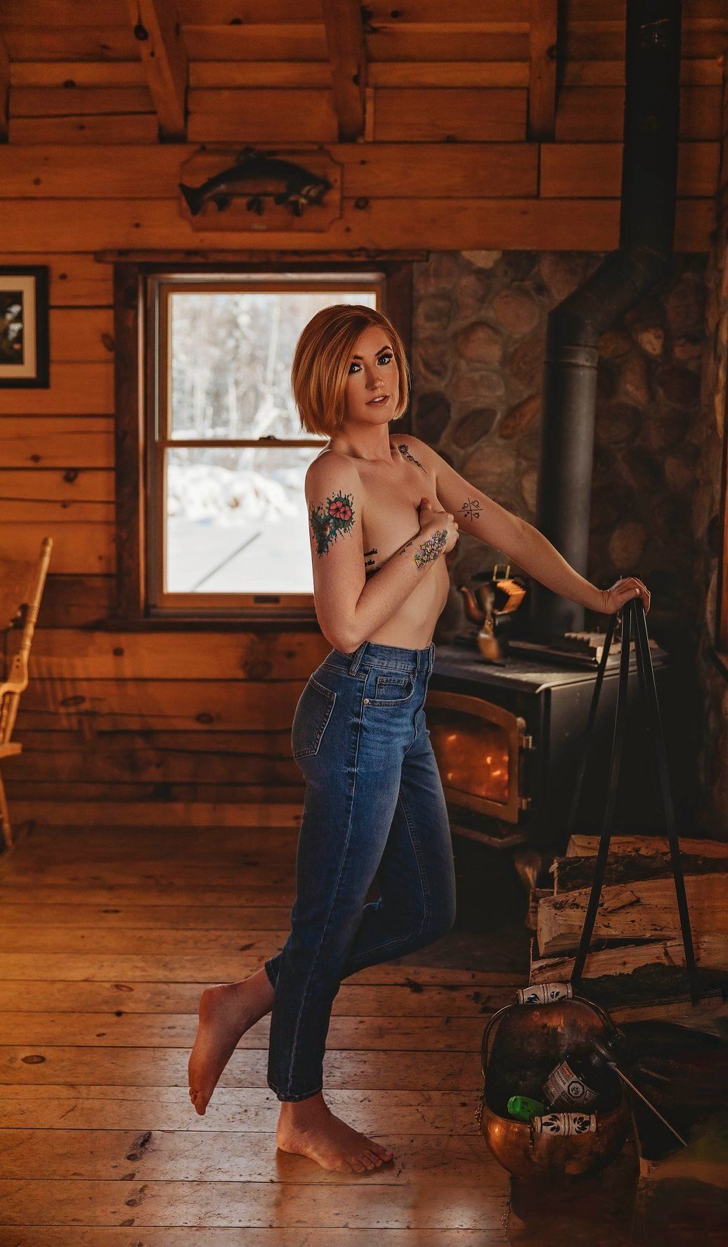 Rustic cabin interior with wood-burning stove, wooden walls and snowy winter scene through window.