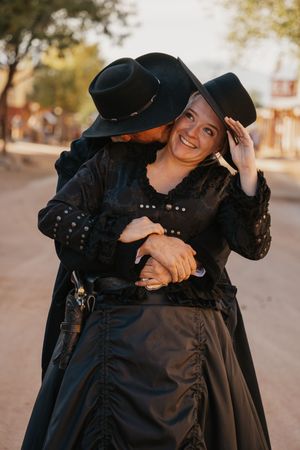 A couple dressed in vintage black clothing embraces and laughs together in warm outdoor lighting.