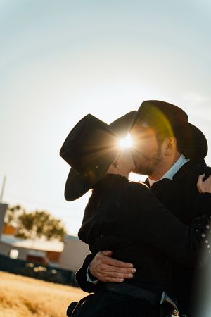 Silhouettes of a romantic couple in cowboy hats embrace against a bright sunset sky.