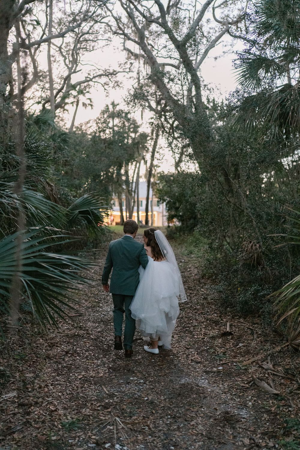 Couple walks down palm-lined forest path at sunset, bride's white dress trailing behind.