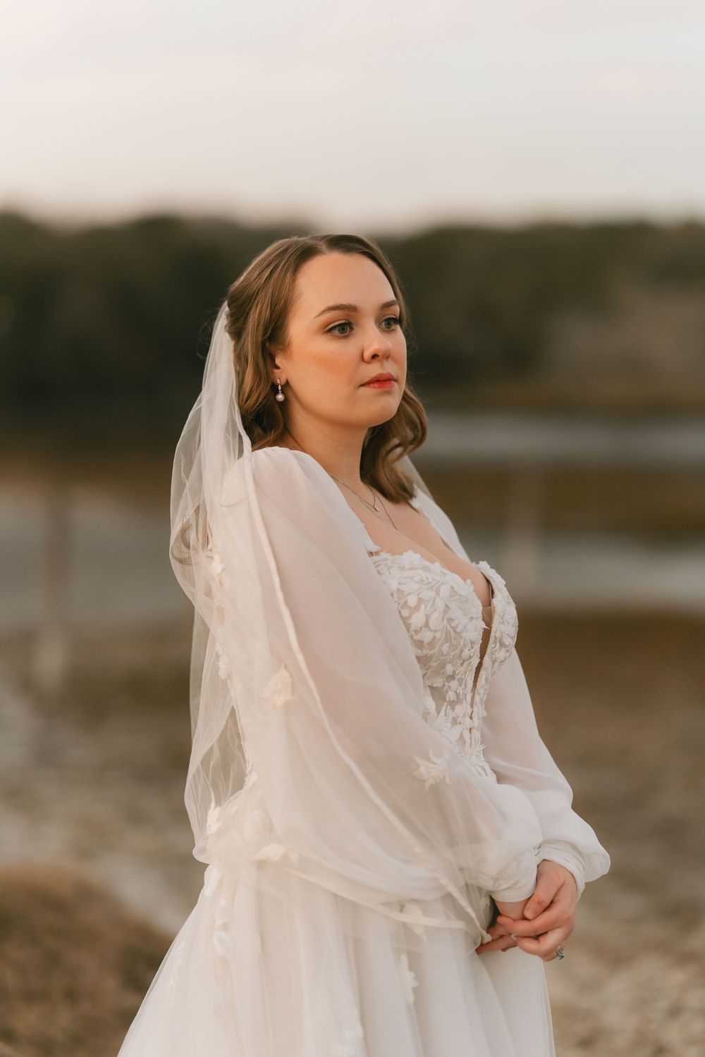 Bride in flowing white dress poses against sunset backdrop near water.
