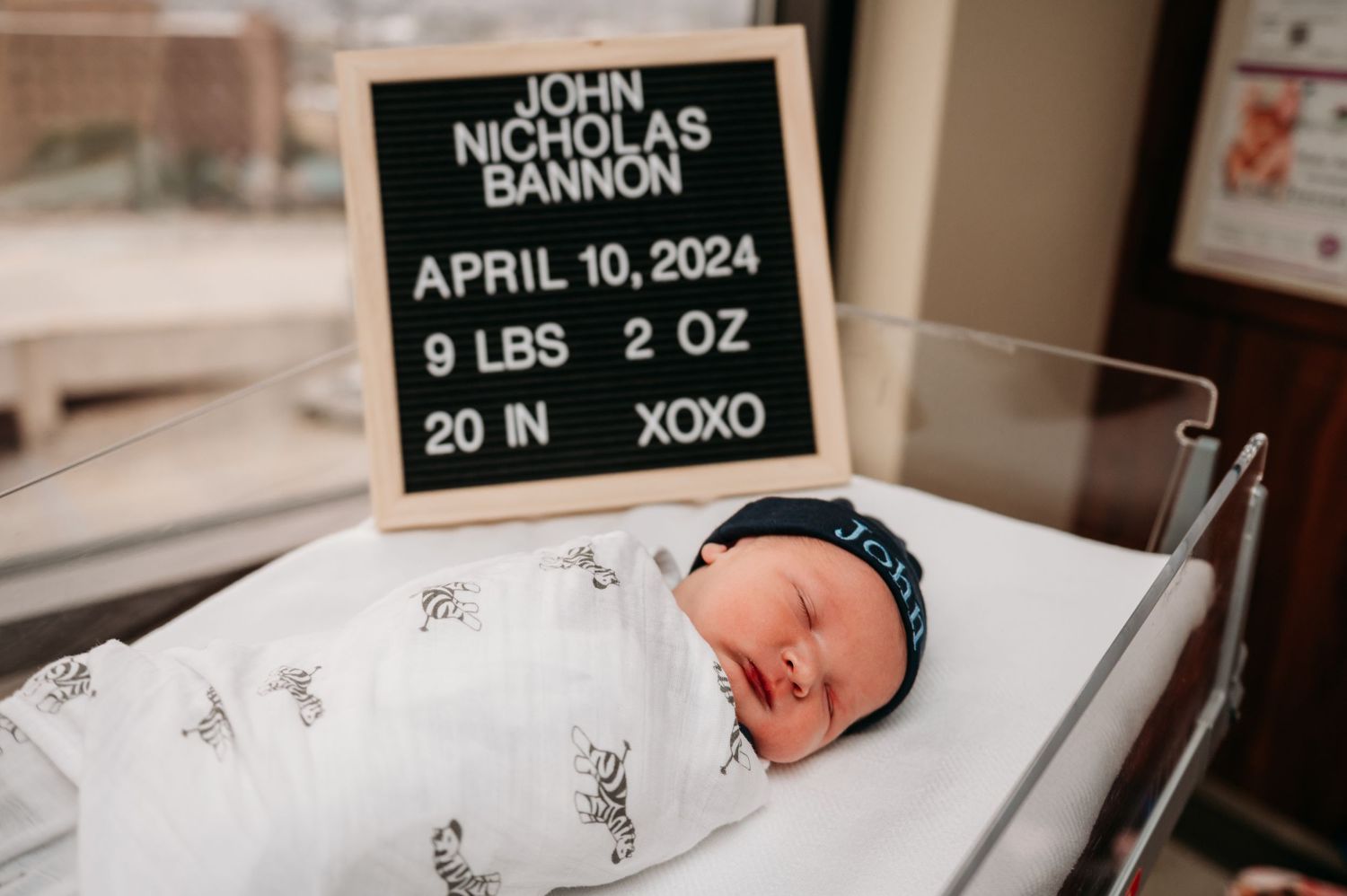 A newborn baby sleeps peacefully in a hospital bassinet with a black letter board displaying birth details above.