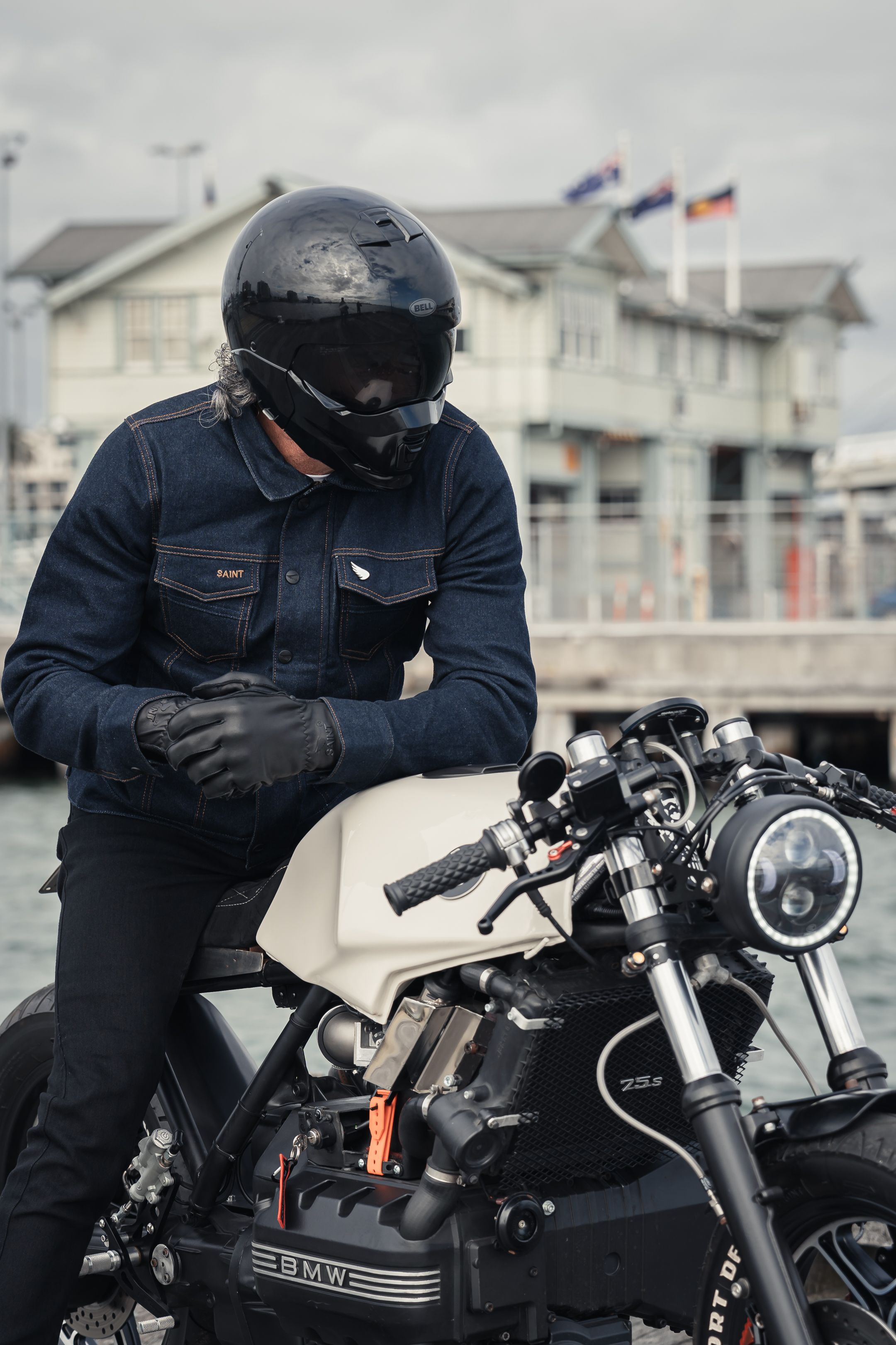 Rider in denim jacket sits on classic BMW motorcycle with houses in background.