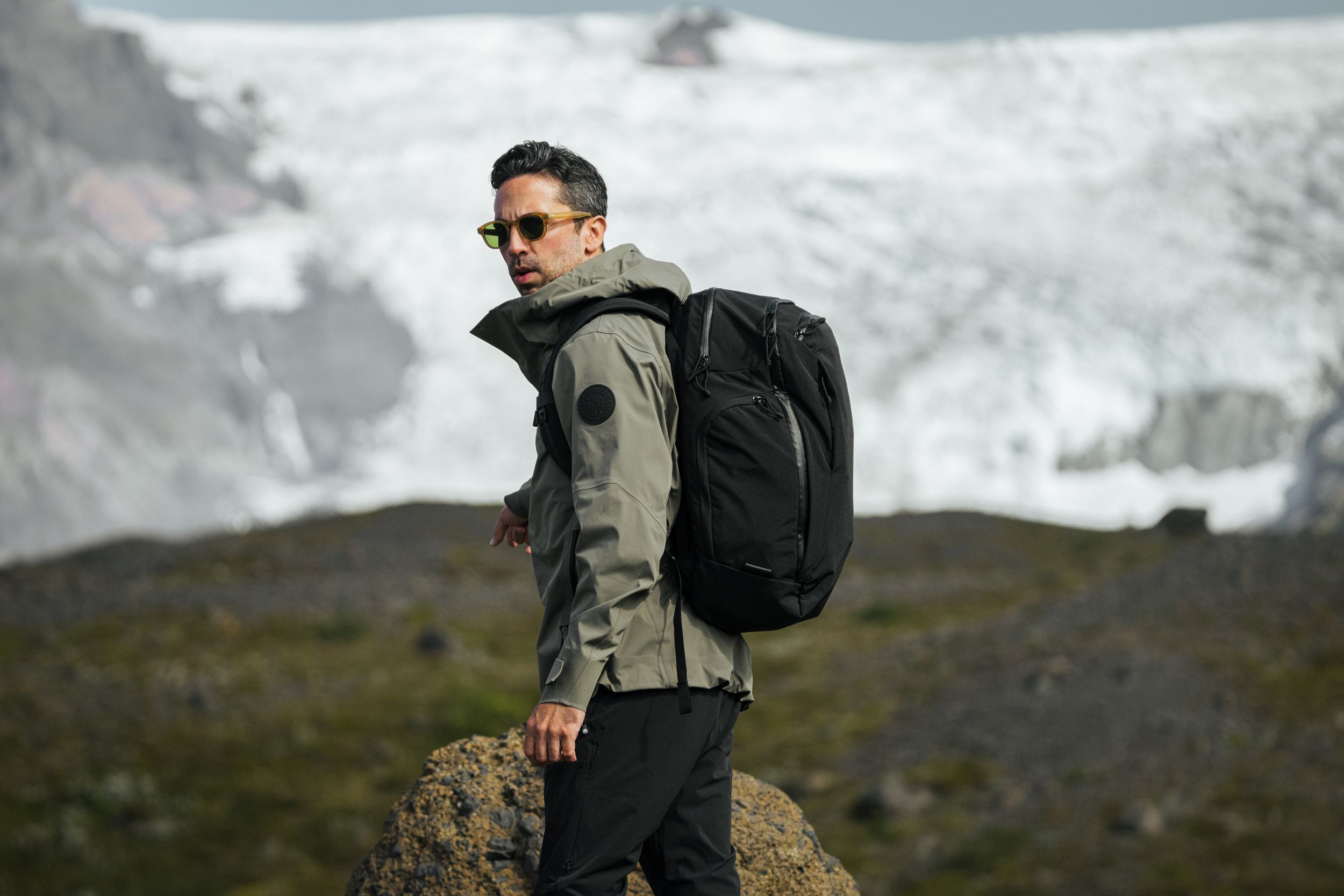 Adventurer with backpack standing on rocky outcrop overlooking glacial landscape.