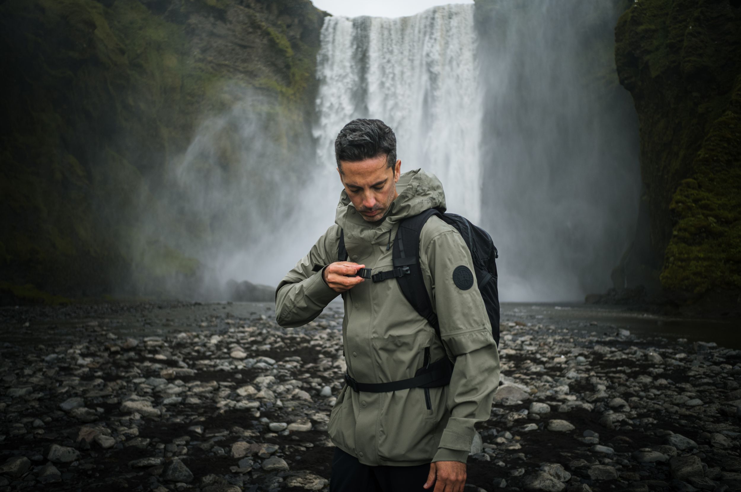 Hiker checking phone while standing at base of powerful waterfall on rocky terrain.