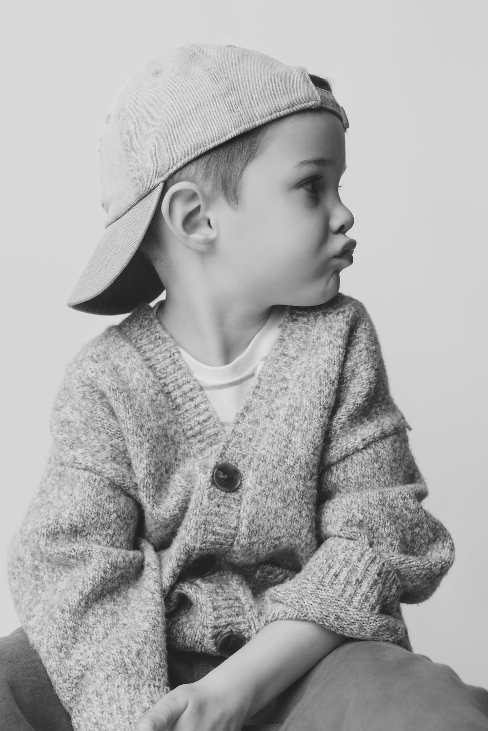 Black and white portrait of a young boy wearing a backward cap and knit cardigan, looking sideways.