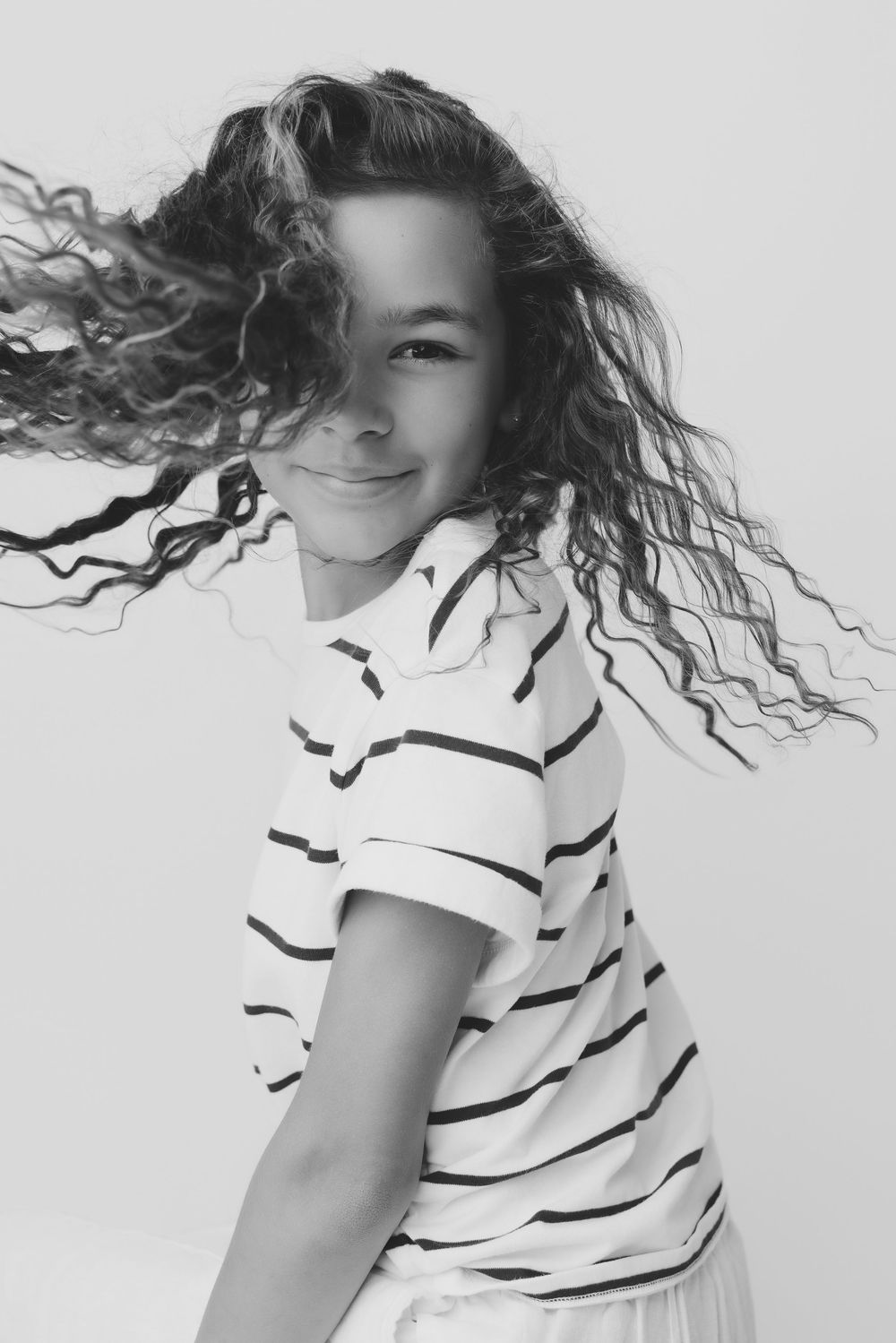 Black and white photo of a smiling child with curly hair flowing, wearing a striped t-shirt.