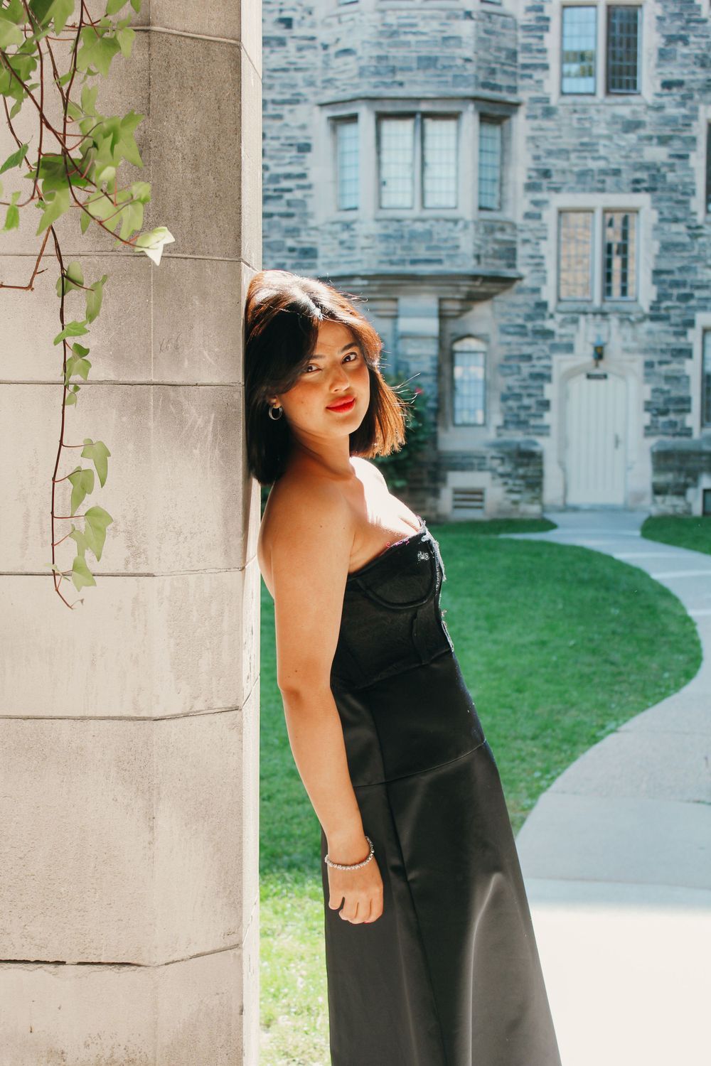 A woman in a black dress poses against a stone pillar with a Gothic building in the background.