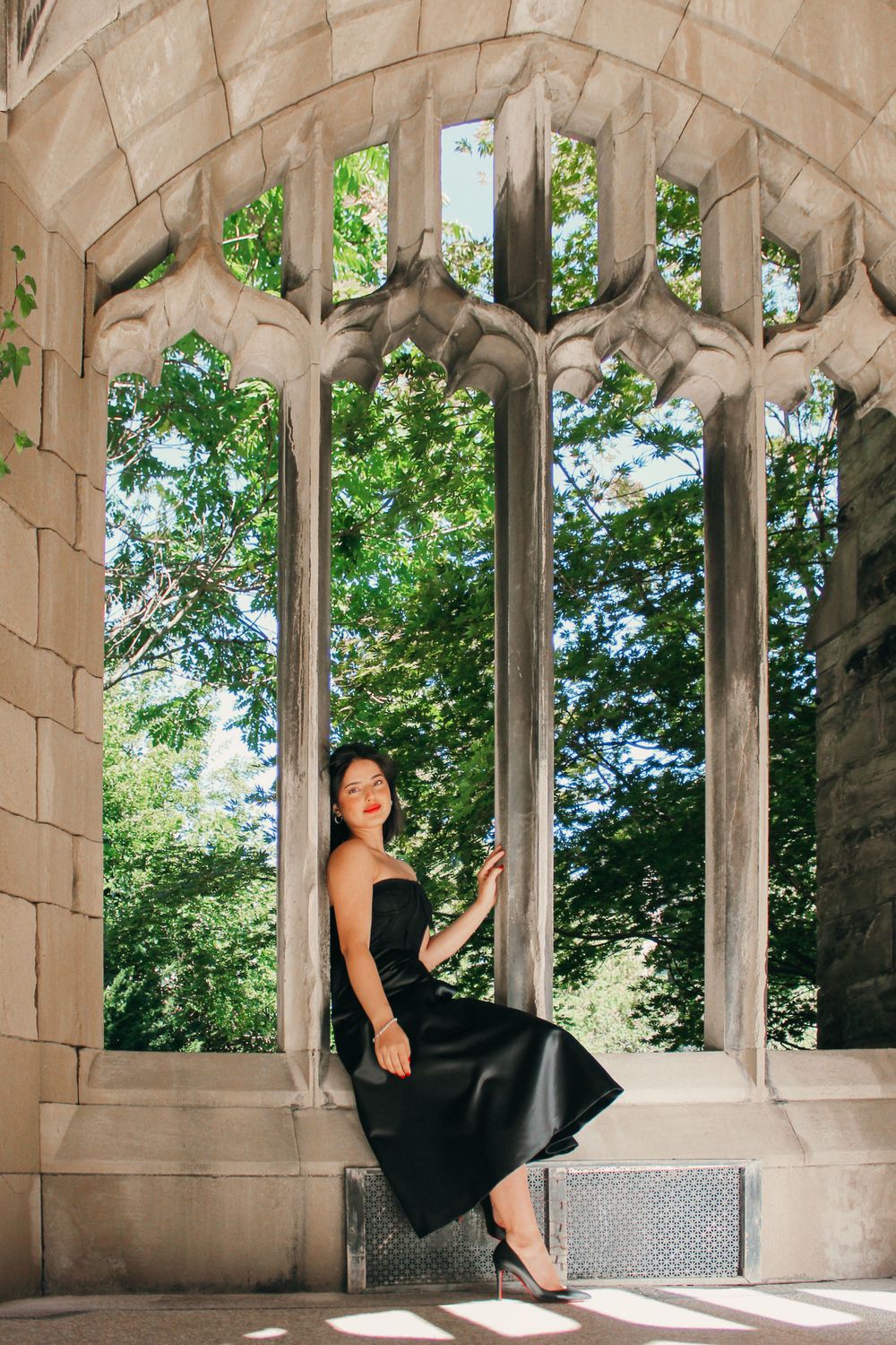 A woman in a black dress poses in a Gothic stone archway framing lush green trees outside.