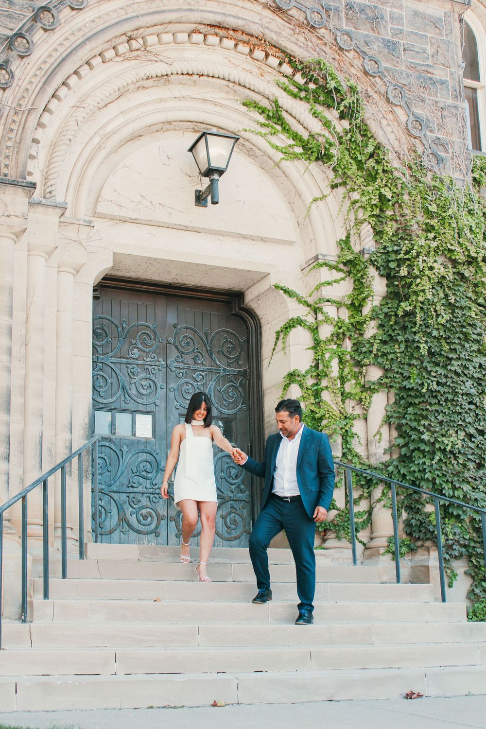 A couple descends stone steps in front of a historic building with ivy and ornate architecture.