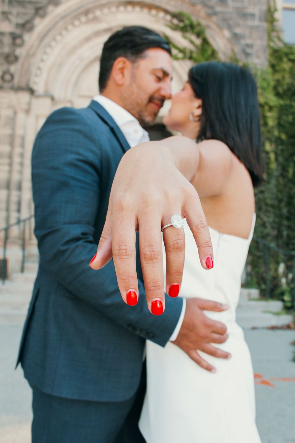Close-up of an engagement ring with red manicured nails against a white dress.