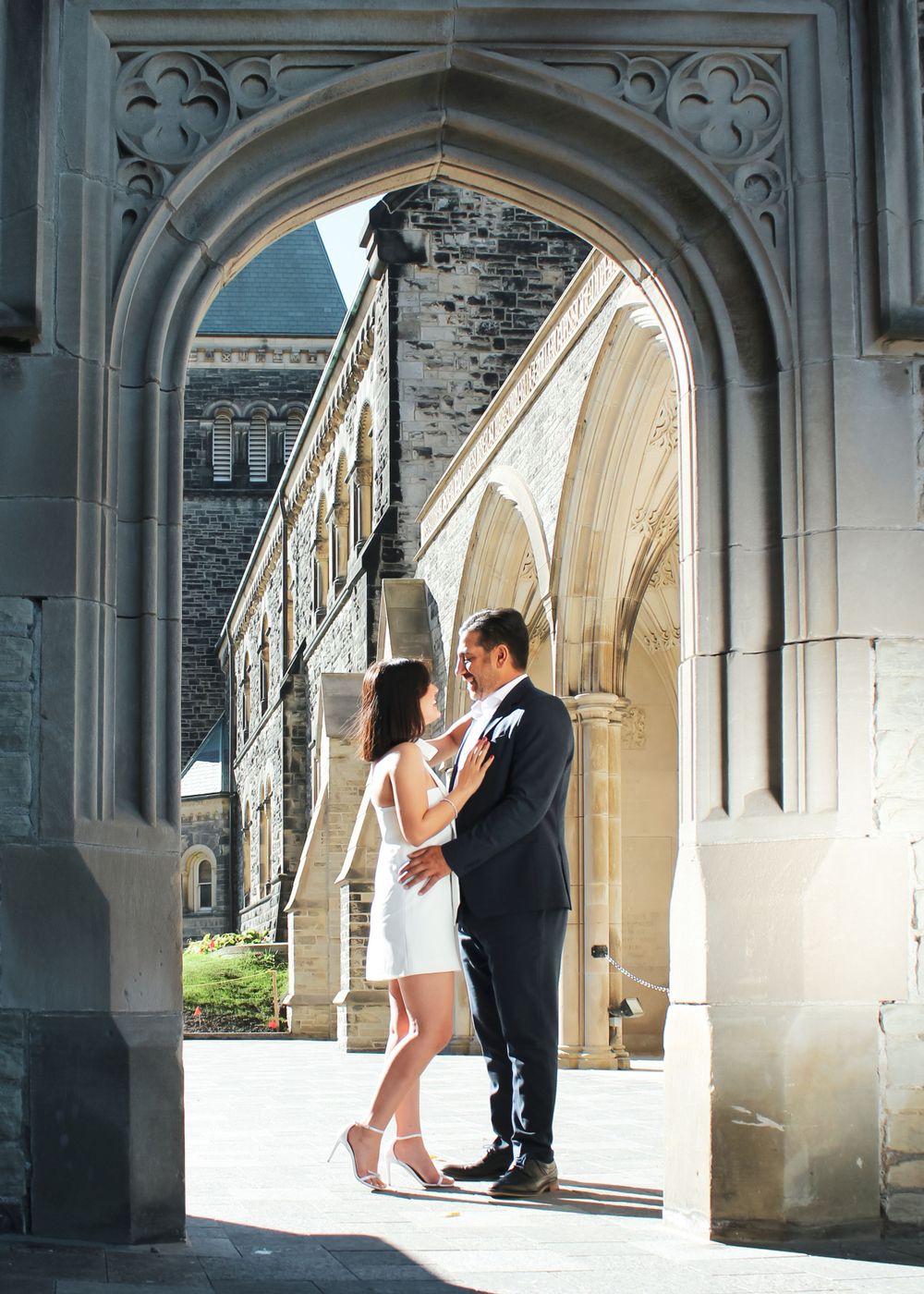 A couple embraces under a Gothic stone archway on a sunny afternoon.
