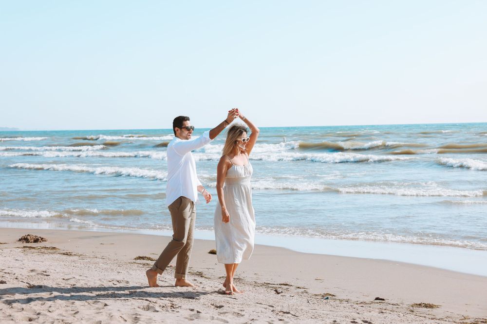 A couple dances joyfully on the beach with ocean waves and blue sky in the background.