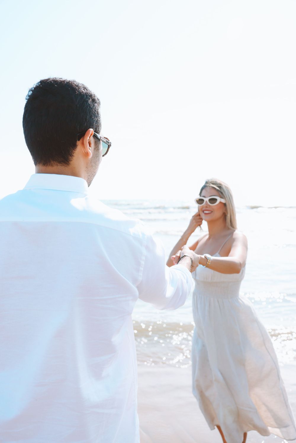 A couple in white clothing dances together on a sunny beach.
