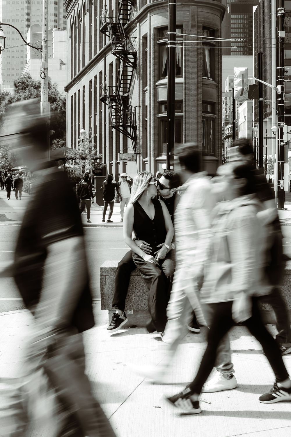 Black and white photo showing motion blur of pedestrians walking past historic building in urban setting.