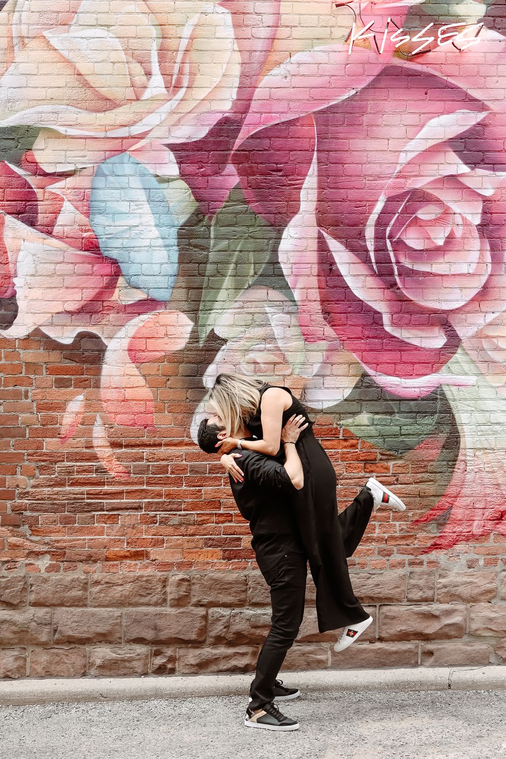 Two people sharing a romantic dance pose in front of a vibrant floral mural on a brick wall.