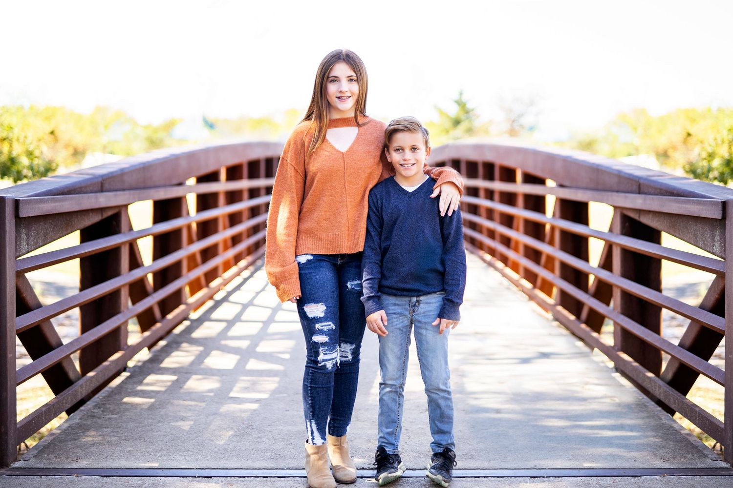 Woman and young child stand together smiling on a wooden bridge during a sunny outdoor photoshoot.