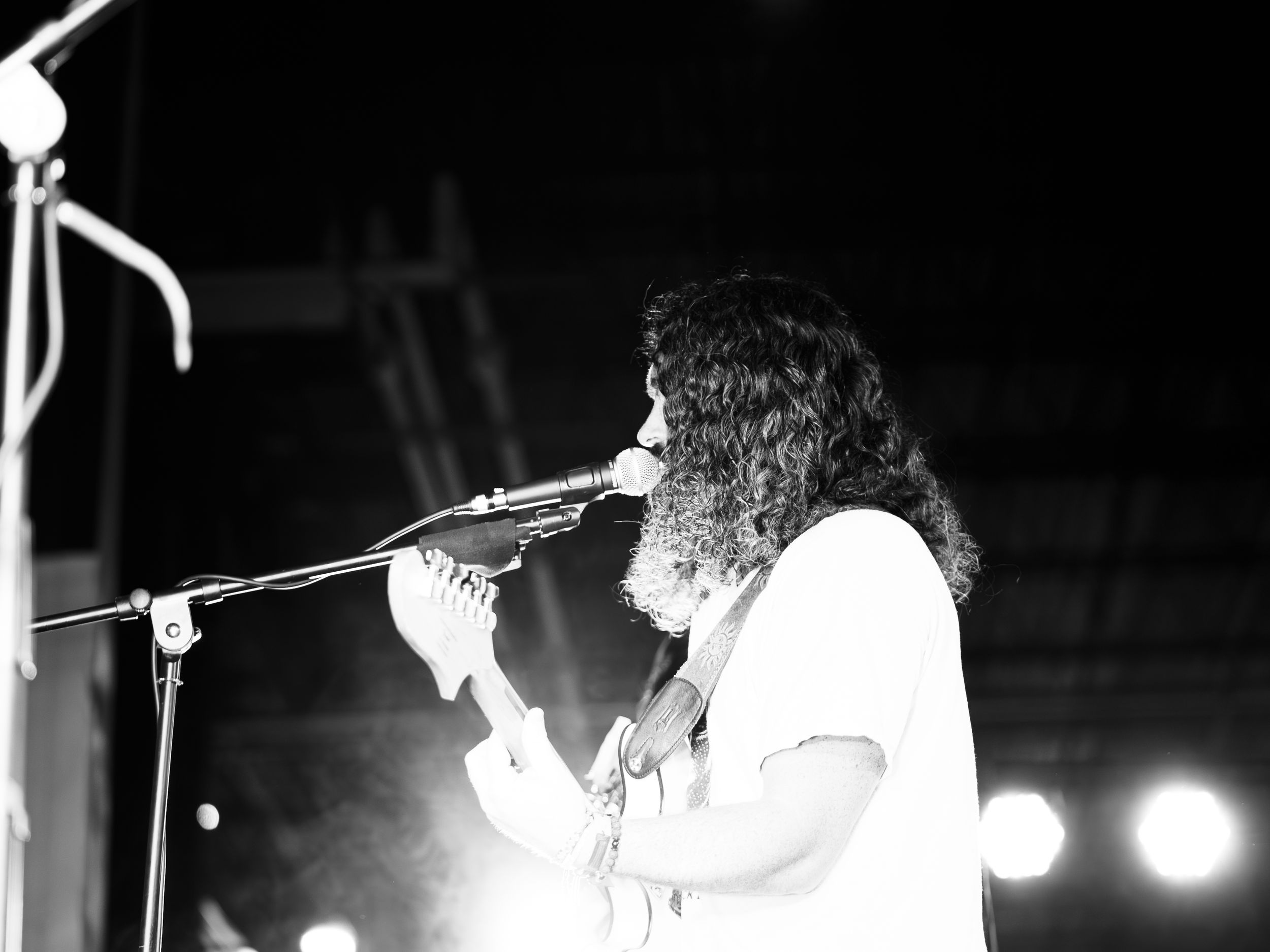 Black and white concert photo of a performer singing into a microphone with curly hair and white shirt.