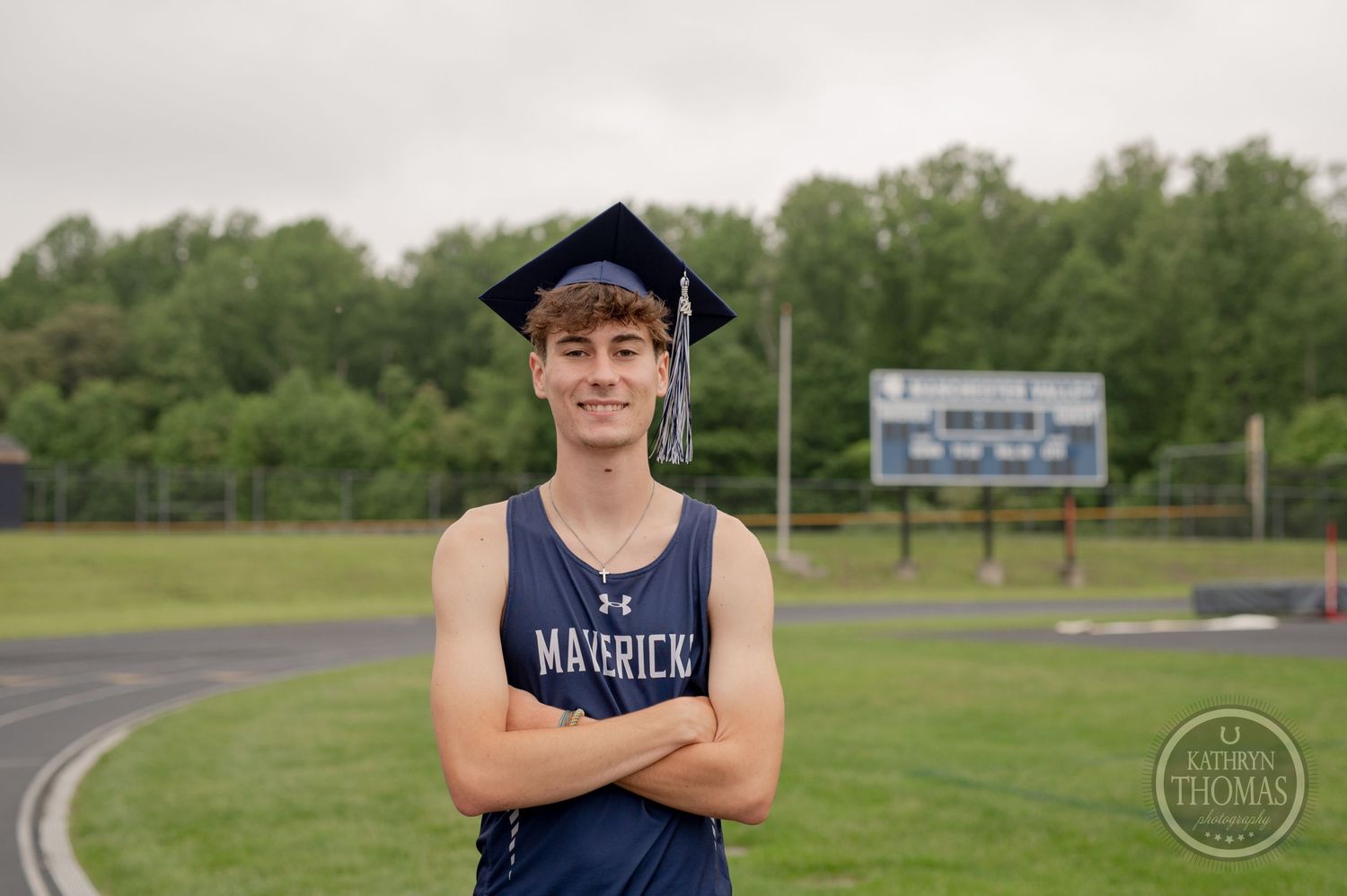 Graduate in navy Mavericks uniform stands with arms crossed on athletic field.