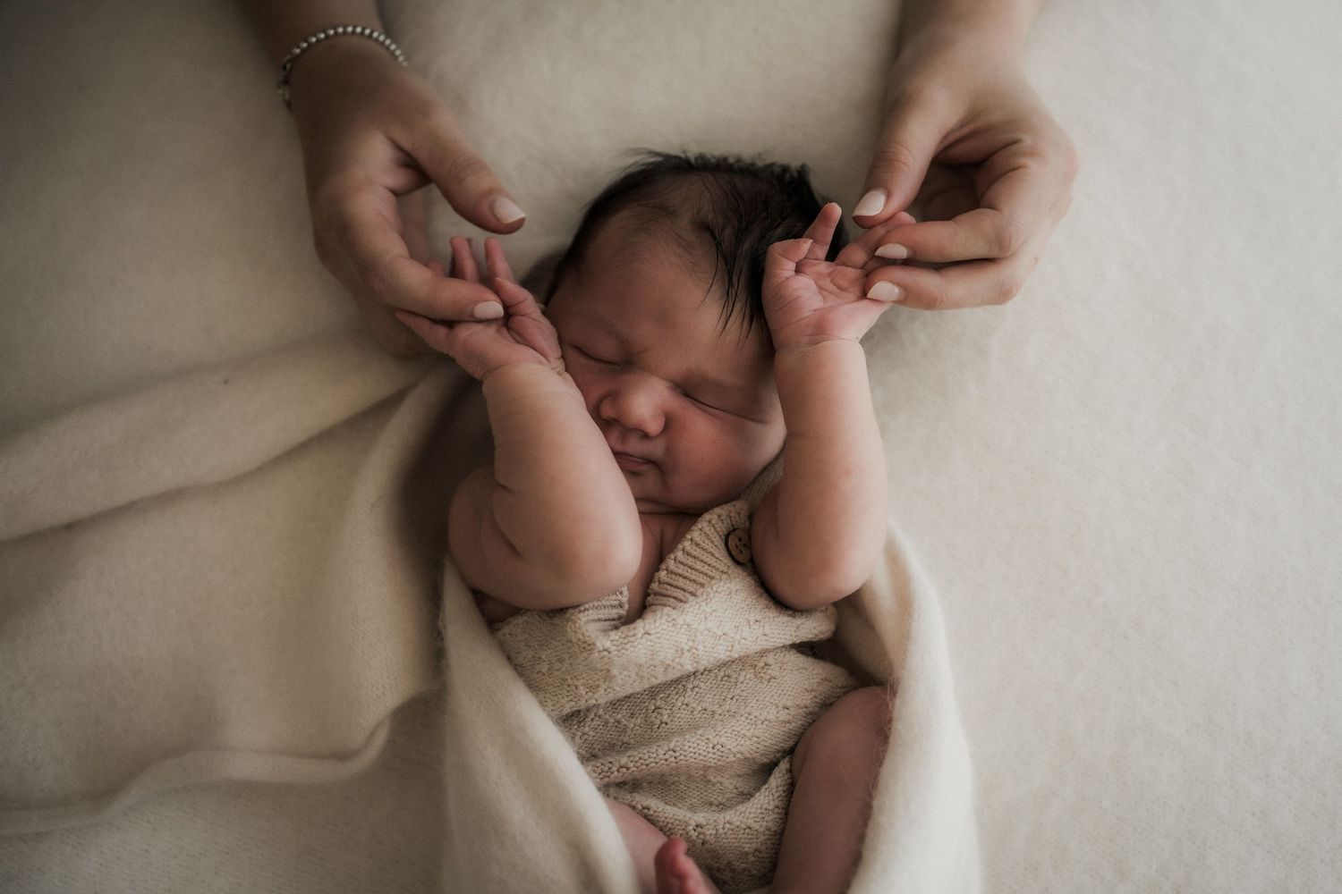 Newborn baby sleeping peacefully wrapped in cream blanket while adult hands gently hold tiny fingers in the Yarra Valley