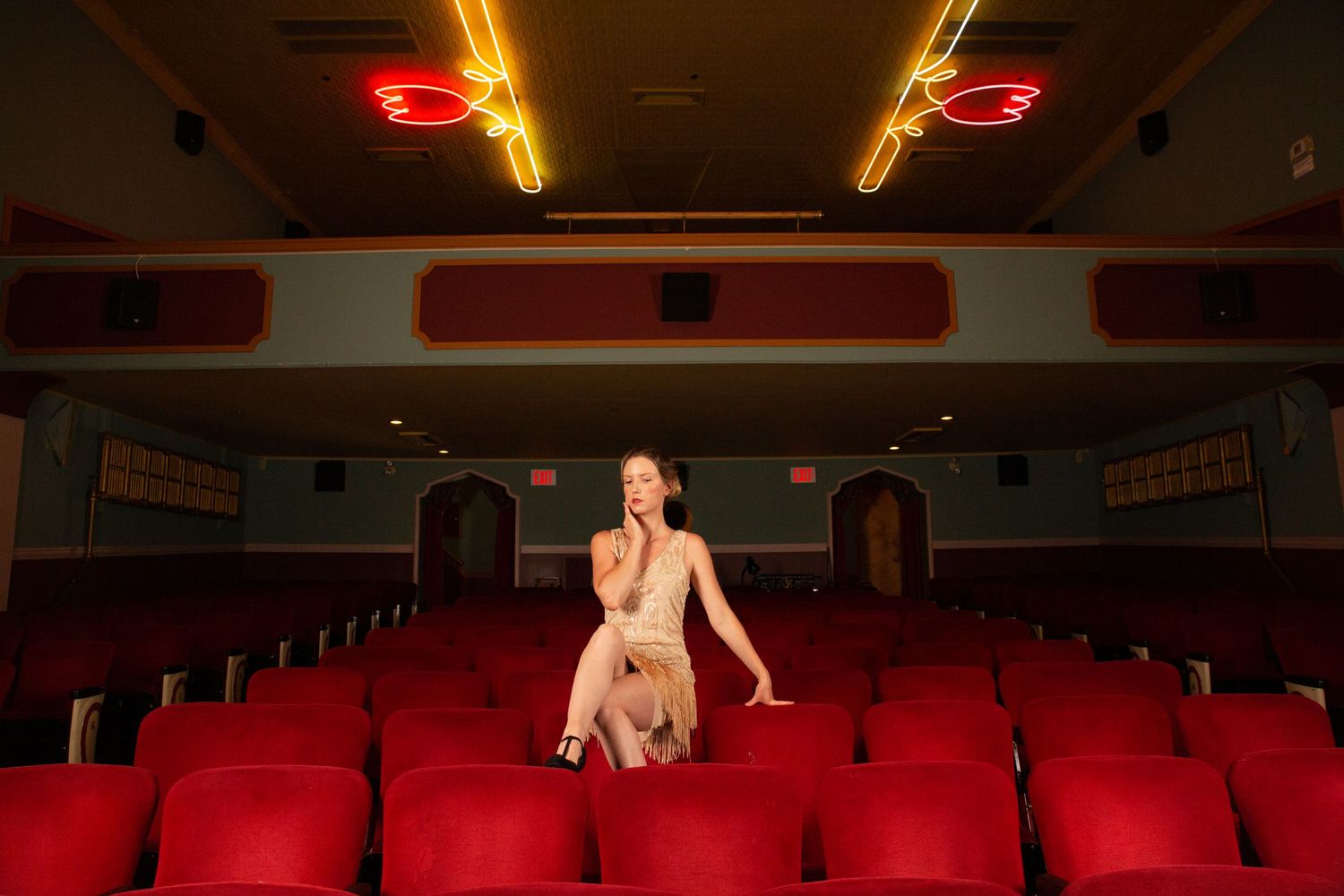 A dancer poses dramatically in an empty theater with red seats and dramatic lighting.