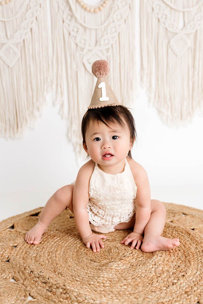 Baby girl in a cream lace romper and birthday hat sits on a jute rug for her first birthday cake smash photo.