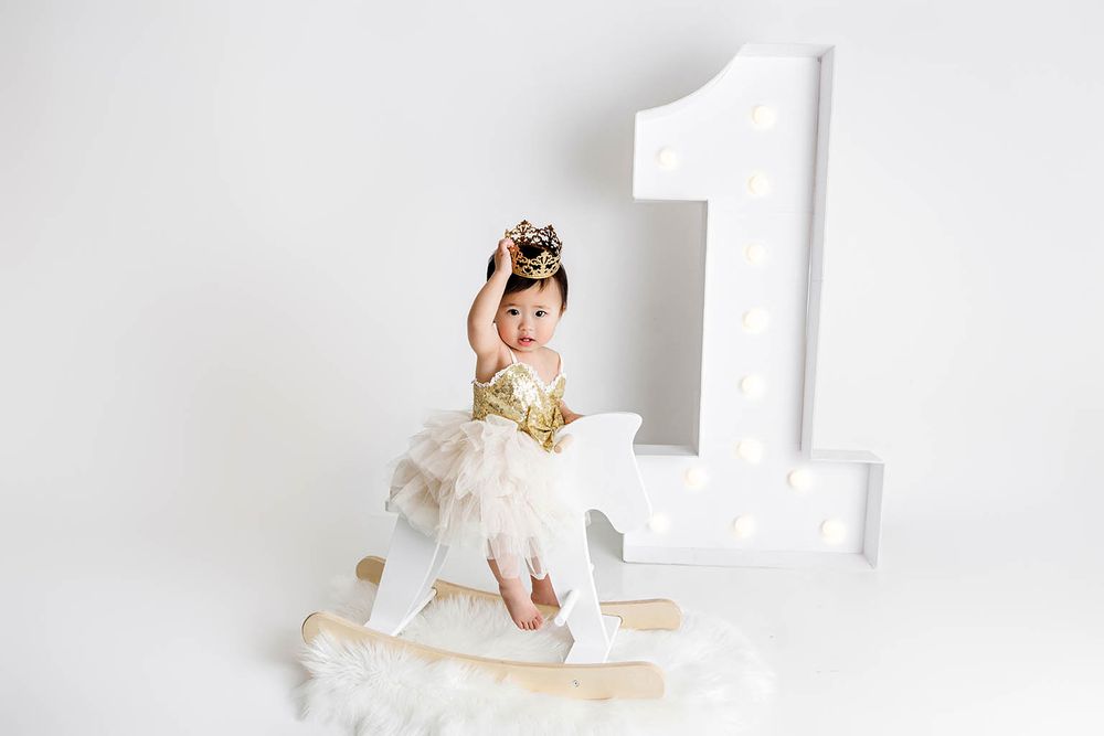 Toddler girl in gold dress sits on rocking horse beside illuminated number 1 for first birthday photo.