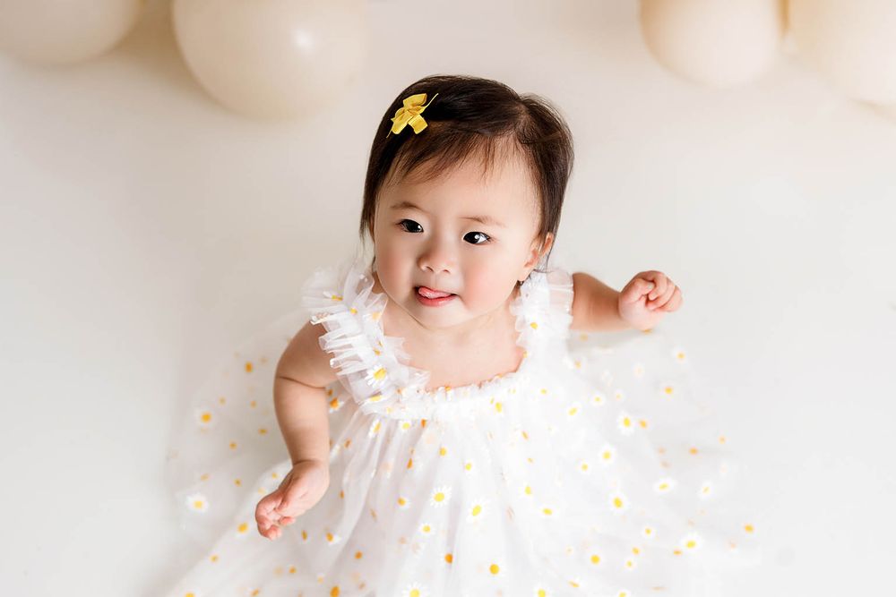 Smiling baby girl in white floral tulle dress with yellow hair bow, sitting among pastel balloons in studio photo.