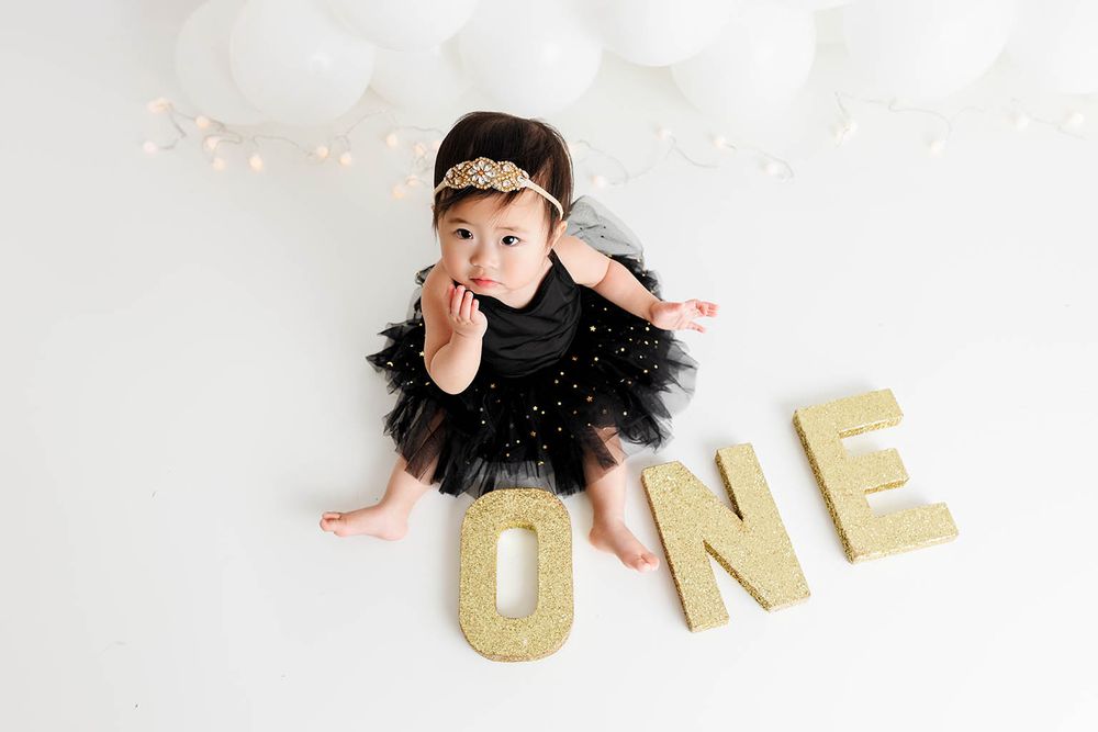 Baby girl in black tutu sits beside gold ONE letters celebrating her first birthday with white balloons.