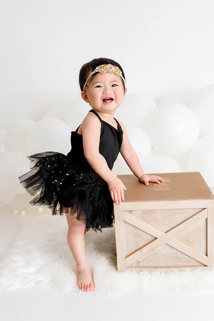 Smiling baby girl in black tutu dress and gold headband standing next to wooden crate on white fur rug.