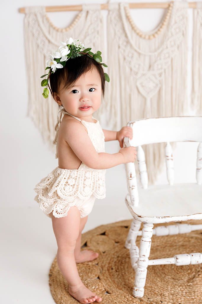 Adorable baby girl in cream lace romper and floral crown stands beside a white chair with macrame backdrop.