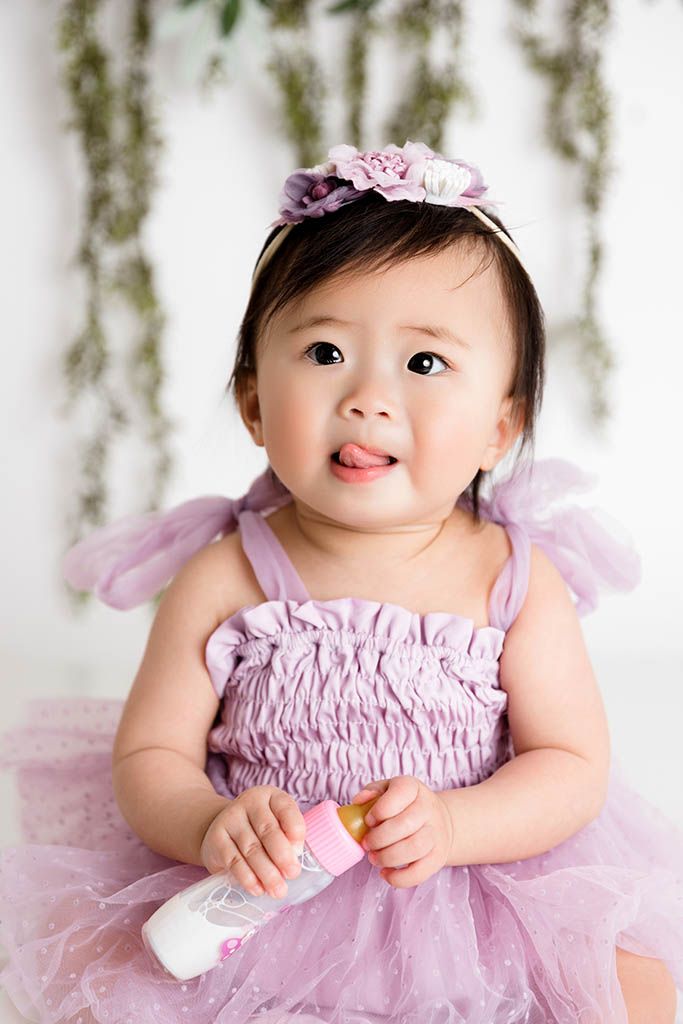 Adorable baby girl in purple tutu dress with floral headband holding a bottle, sitting against white backdrop.