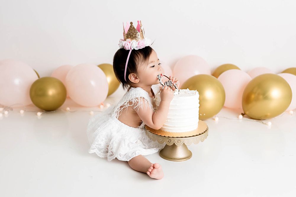Baby girl in white dress and pink crown smashes first birthday cake surrounded by pink and gold balloons.