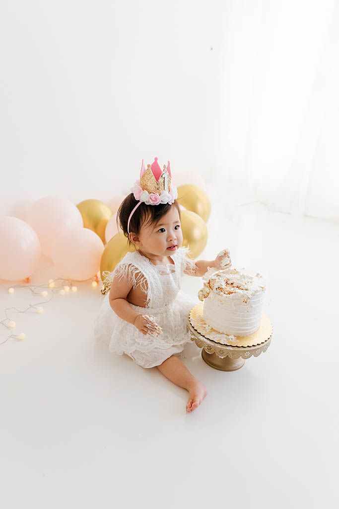 Baby girl in white lace dress and pink crown smashes first birthday cake surrounded by pink and gold balloons.