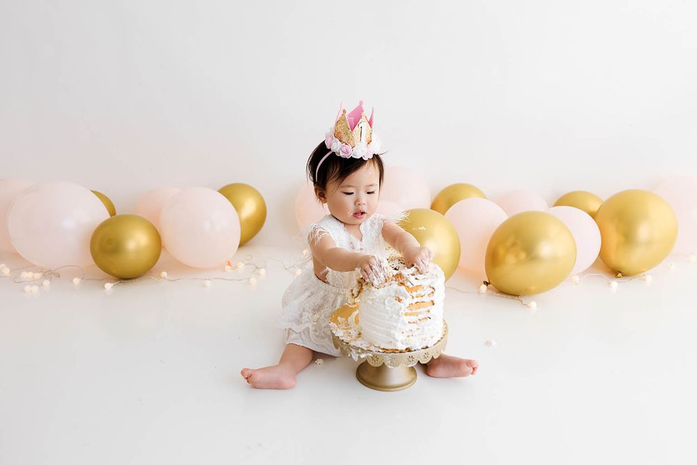 Baby girl in white dress and pink crown smashes first birthday cake surrounded by pink and gold balloons.