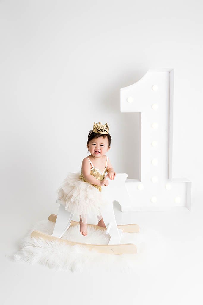 Baby girl wearing gold crown and tutu sits on rocking horse beside illuminated number one.