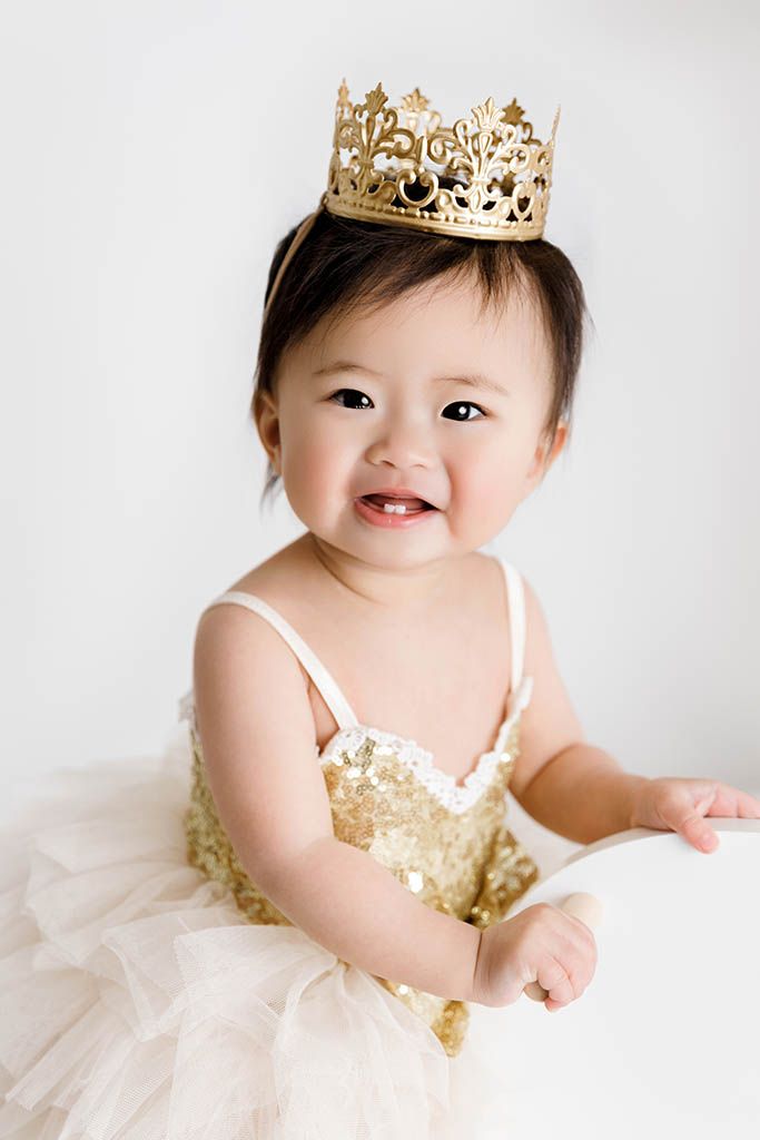 Smiling baby girl in gold sequin dress and crown celebrates her first birthday portrait.