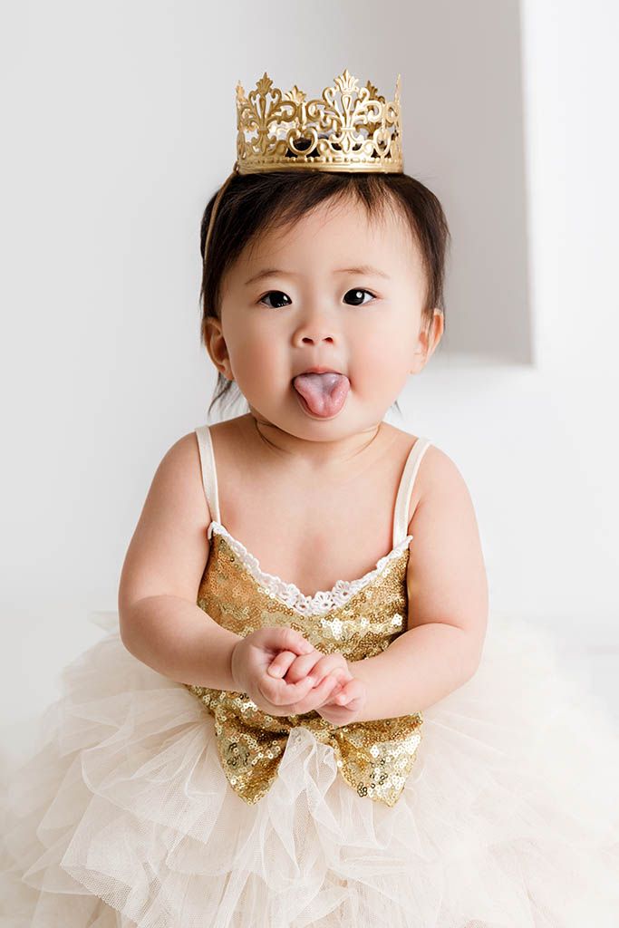 Adorable baby girl in gold sequin dress and crown sticking out her tongue, perfect for first birthday photos.