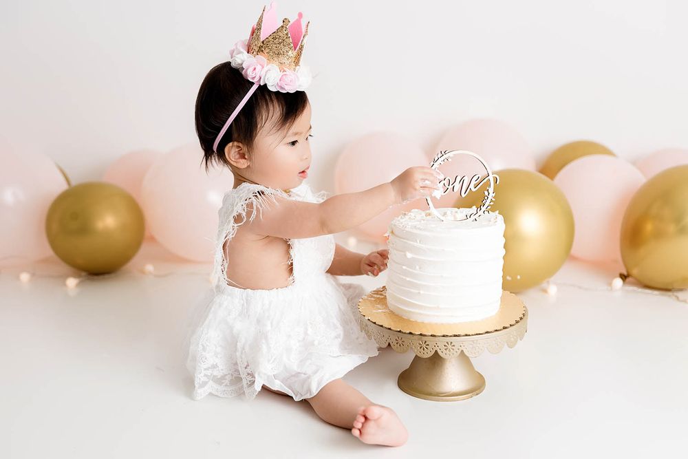 Baby girl in white dress touches first birthday cake on gold stand, pink and gold balloons behind her.