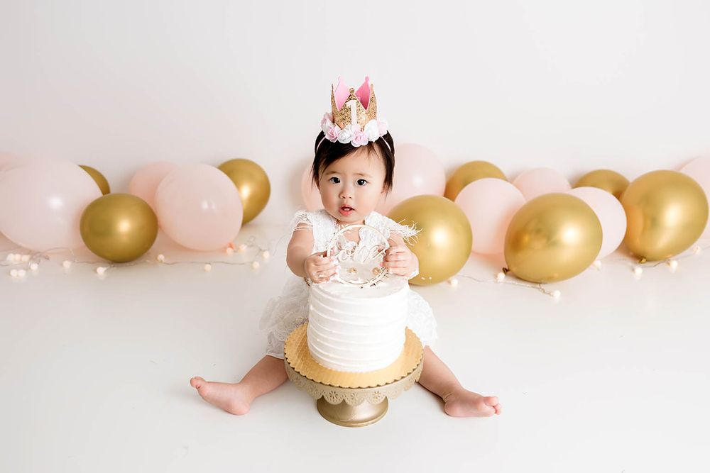 Baby girl in white dress and crown sits with first birthday smash cake surrounded by pink and gold balloons.